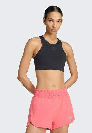 Atletka o ciemnych włosach, ubrana w czarny bezrękawnik typu crop top i różowe szorty z elastycznym pasem, stoi z rękami za plecami.