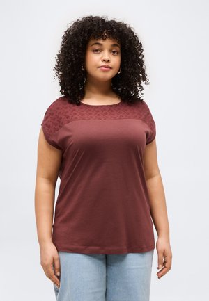 Kvinde med krøllet hår iført en vinrød bluse med korte ærmer og blonder samt lyseblå jeans, stående foran en ensfarvet baggrund.