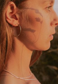 Silberne Creolen, schlanker geschichteter Halsreifen, warme Hauttöne, welliges Haar, sanfte Schatten am Hals. Der Hintergrund zeigt unscharfe natürliche Elemente.
