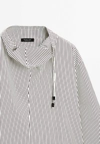 Weißes Hemd mit schwarzen vertikalen Streifen, ausgestattet mit einem umgelegten Kragen und einer dekorativen Schnur mit schwarzer Hardware zur Verstellung.