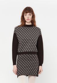 Donna con capelli rossi che indossa un maglione a righe marroni e una gonna abbinata, in piedi davanti a uno sfondo bianco uniforme.