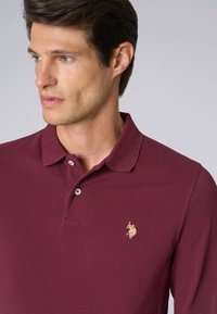 Polo shirt bordeaux con colletto classico, pattina con due bottoni e un piccolo logo ricamato in oro sul lato sinistro del petto. Materiale in cotone, finitura strutturata.
