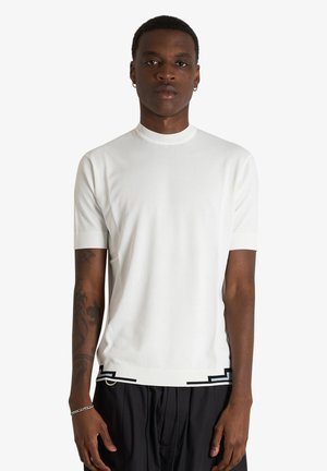 Uomo con pelle scura e capelli corti indossa una camicia bianca a maniche corte con pantaloni neri, in piedi su uno sfondo bianco uniforme.
