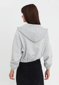 Kobieta z brązowymi włosami, ubrana w jasnoszary krótką bluzę z kapturem i czarną spódnicę dżinsową w wysokim stanzie, zwrócona plecami na białym tle.