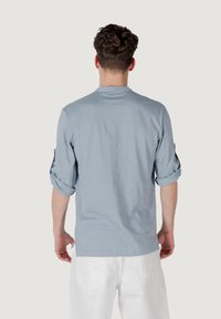 Maglietta azzurra chiara a maniche lunghe con maniche arrotolate, colletto rotondo e tessuto morbido e texture. Indossata con pantaloni bianchi.