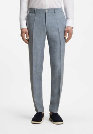 Man draagt lichtgrijze op maat gemaakte broek, wit overhemd en donkere instapschoenen, staand tegen een effen witte achtergrond.