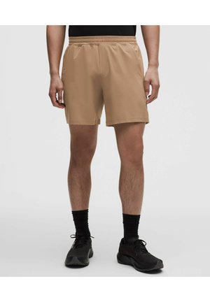 Hombre con pantalones cortos deportivos de color beige, calcetines negros y zapatillas de running negras, de pie frente a un fondo liso.