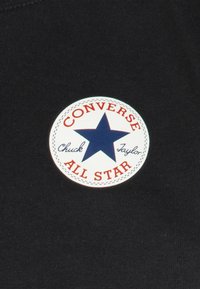 Toppa circolare bianca con una stella blu scura, testo rosso con la dicitura "CONVERSE" e "ALL STAR", su uno sfondo di tessuto nero.