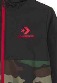 Chaqueta negra con cremallera roja, logo rojo de Converse con una estrella y una flecha, y patrón de camuflaje en la parte inferior en verde, marrón y beige.