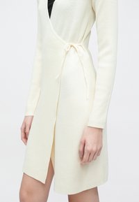 Ribbstrikket kjole i kremfarge med omslagsdesign, lange ermer, sideslå med knyting og høyt splitt på venstre side.