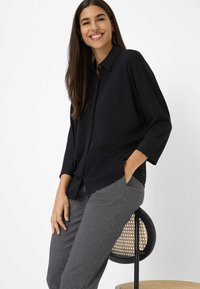 Zwarte button-up shirt met een losse pasvorm en driekwartmouwen, gecombineerd met grijs geruite broek. Model staat casual op een stoel.