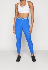 Niebieskie legginsy sportowe z wysokim stanem z napisem "NIKE PRO" w białym kolorze, w połączeniu z białym stanikiem sportowym i czarnymi butami.