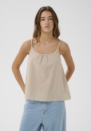 Junge Frau mit langen braunen Haaren trägt ein beige-weiß gestreiftes ärmelloses Oberteil und einen hellblauen Jeansrock, posiert vor einfachem Hintergrund.