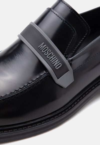 Mocassin en cuir noir brillant avec des détails cousus et une sangle grise sur le dessus embossée du logo "MOSCHINO".