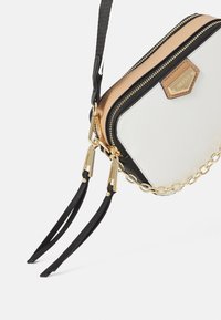 Kompaktowa torebka z białym, teksturowanym korpusem, czarnymi akcentami i złotym suwakiem. Posiada łańcuszkowy pasek oraz skórzany, beżowy detal.