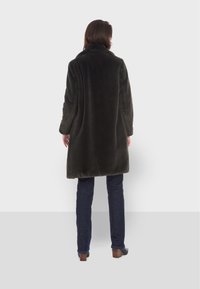 Manteau en fausse fourrure vert foncé avec un col haut, longueur genoux et manches longues, porté sur un jean foncé et des talons. Texture lisse avec une coupe décontractée.