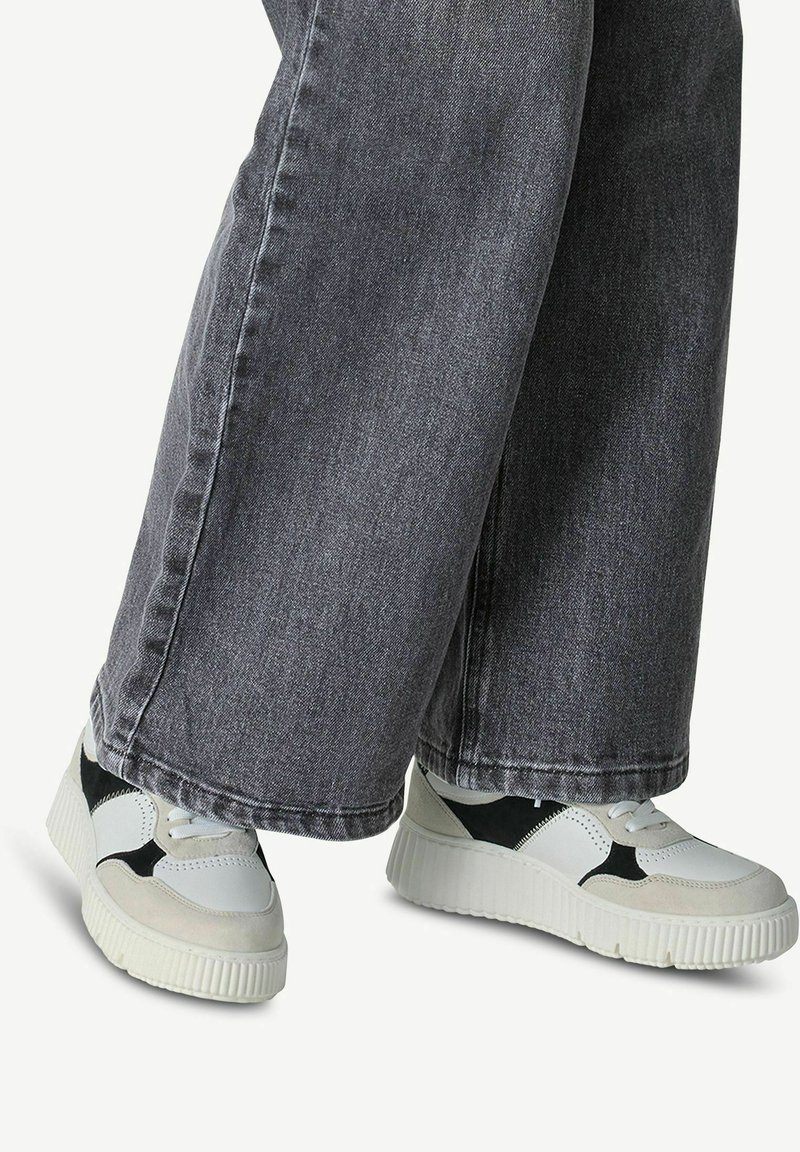 Sneakers bianchi e neri con accenti texturizzati, caratterizzati da una suola spessa e un mix di materiali, abbinati a jeans grigi a gamba larga.