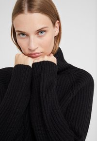 Black knitted sweater with a high collar, featuring ribbed texture and long sleeves, hands clasped at the neck, against a white backdrop.