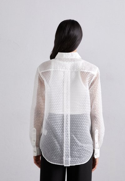 Camicia bianca traslucida con motivo a griglia testurizzato, dotata di colletto classico e maniche lunghe, indossata sopra pantaloni scuri.
