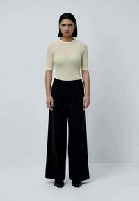 Γυναίκα με ίσια μαύρα μαλλιά, φορώντας ένα μπεζ πλεκτό μπλουζάκι κοντό μανίκι και μαύρο παντελόνι με ψηλή μέση και φαρδιά γραμμή, που στέκεται σε ουδέτερο γκρίζο φόντο.