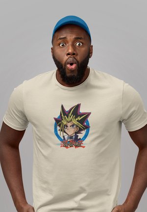 T-shirt beige avec un graphisme d'un personnage aux cheveux blancs et noirs, yeux bleus, entouré d'un emblème coloré, imprimé sur un tissu doux.