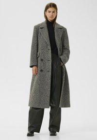 Abrigo cruzado en un patrón de espiga en blanco y negro, con cuello de muesca, bolsillos laterales y un diseño hasta la rodilla.
