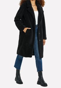 Manteau noir et duveteux avec une texture en shearling, manches longues, col à revers, poches avant et fermeture par boutons. Porté sur un haut blanc et un jean.