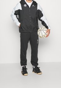Sort vindjakke med grå ærmer, NBA-logo og lynlås. Sorte træningsbukser med hvidt Nike-logo. Sorte sneakers med gummisåler.