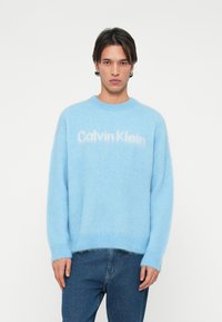 Világoskék pulóver puha, bolyhos anyagból, kerek nyakkivágással. Az elején a "Calvin Klein" felirat látható, egy kicsit világosabb árnyalatban.