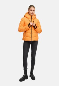 Orange puffjacka med huva, svarta accenter på ärmslut och dragkedja, quiltad textur, ihopparad med svarta jeans och kraftiga svarta kängor.