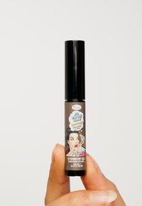 Gel à sourcils dans un tube noir slim avec un design de dessin animé sur l'étiquette. Fond marron, texte clair et ingrédients notés. Taille : 3,8 ml.