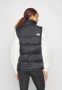 Μαύρο γιλέκο με ψηλό λαιμό, με επλαιωμένο υφή και λογότυπο στον αριστερό ώμο. Φορεμένο πάνω από λευκό μπλουζάκι με μακριά μανίκια.