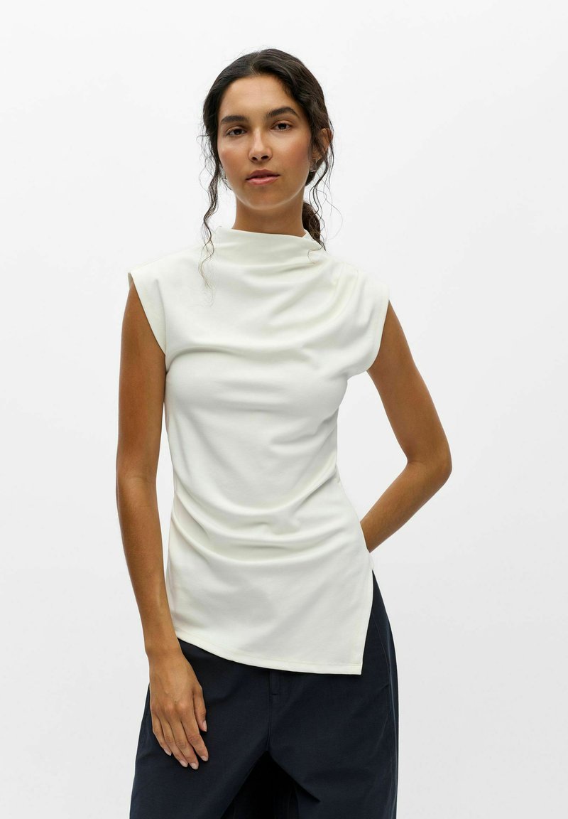 Jeune femme aux cheveux foncés attachés en queue de cheval basse porte un haut blanc sans manches à col montant et un pantalon foncé sur un fond blanc uni.