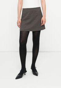 Ženska v sivi mini krilu, črnih neprozornih nogavicah in črnih slingback kitten heels s koničastim nosom, stoji ob beli steni.
