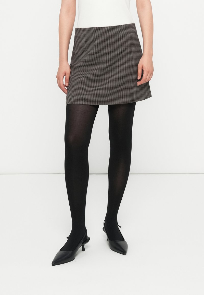 Ženska v sivi mini krilu, črnih neprozornih nogavicah in črnih slingback kitten heels s koničastim nosom, stoji ob beli steni.