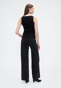 Čierny rebrovaný tielko s výstrihom do V, skombinované so širokými čiernymi džínsami. Modelka zobrazená zozadu, zdôrazňujúca strih oblečenia.