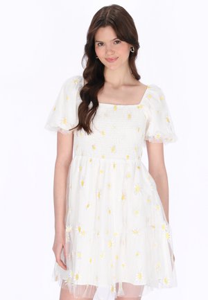 Ung kvinde med langt brunt hår iført en hvid kjole med pufærmer og gul margueritbroderi, der smiler let.