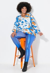 Top a motivo blu e arancione con simbolo della pace, leggings blu lucidi e stivali neri con dettagli borchiati, seduta su uno sgabello arancione.