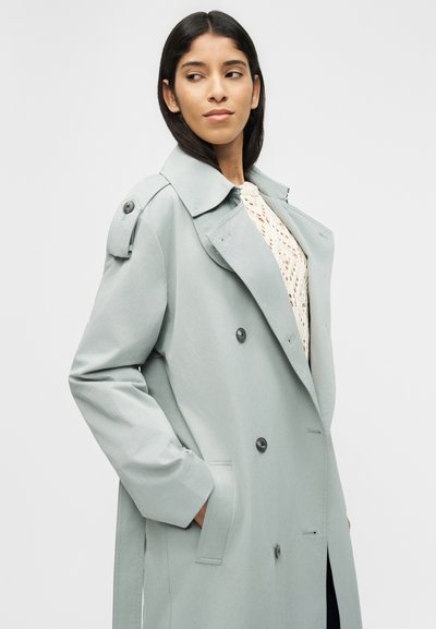 Γυναίκα που φοράει ανοιχτό γκρι διπλ ής όψης καμπαρντίνα με πλεκτό εκρού πουλόβερ, κοιτώντας προς τα πλάγια με τα χέρια στις τσέπες.