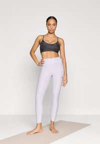 Czarny pasiaste stanik sportowy, jasnofioletowe legginsy z wysokim stanem. Oba wykonane z rozciągliwego, gładkiego materiału. Na boso na jasnobeżowej macie.