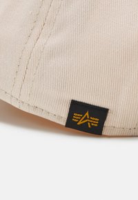 Beige sapka textúrált, bordázott anyagból. Fekete címkével rendelkezik, sárga logóval és varrással az illesztéseknél. Gömbölyű alakú, napellenző nélkül.