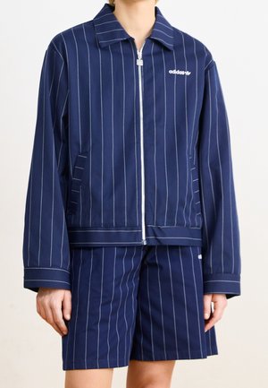 Osoba nosící tmavě modrou proužkovanou bundu na zip s límečkem a odpovídající krátké kalhoty po kolena, na bundě je logo Adidas na hrudi.