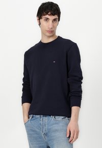Navy blue sweatshirt made of cotton, featuring a crew neck and long sleeves. Small logo accent on the chest, paired with light blue jeans.