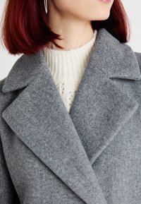 Gray felt coat with a wide lapel, shown over a textured cream sweater. The coat's fabric appears soft and warm.