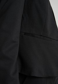 Blazer noir sur mesure en tissu lisse, épaules structurées et une fente arrière unique. Lignes épurées et design minimaliste.