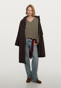 Lång, mörkbrun trenchcoat bärs över en grå stickad V-ringad tröja. I kombination med högmidjade blå jeans och bruna slip-on skor.