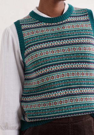 Osoba ubrana w dzianinową kamizelkę w kolorach morskim, białym i rdzawym, na białej bluzce z długim rękawem, spod której widoczne są brązowe spodnie sztruksowe.