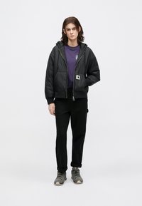 Schwarze gesteppte Jacke mit Kapuze, mit einem Reißverschluss und Vordertaschen. Über einem lila T-Shirt getragen, kombiniert mit schwarzen Hosen und grauen Schuhen.