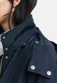 Mörkblå jacka med hög krage, prydd med silverfärgade tryckknappar, slätt tyg och en strukturerad design. Accenter på kragen och axlarna är synliga.
