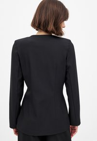 Schwarzer maßgeschneiderter Blazer mit langen Ärmeln, glatter Textur, tailliertem Schnitt und einem leicht längeren Rückensaum, ohne sichtbare Hardware oder Akzente.